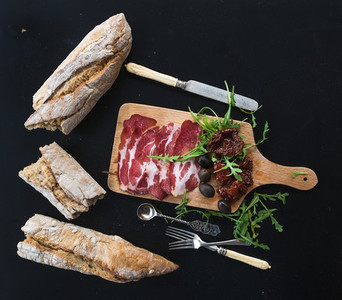 Wine appetizer set vintage dinnerware french baguette broken into pieces dried tomatoes olives smoked meat and arugula on rustic wooden board over dark background