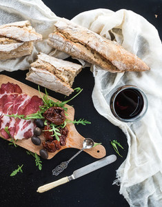 Wine appetizer set vintage dinnerware french baguette broken into pieces dried tomatoes olives smoked meat and arugula on rustic wooden board over dark background