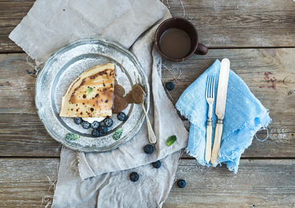 Thin pancake or crepe with fresh blueberry cream mint and salty caramel sauce in vintage metal plate over rustic wooden backdrop