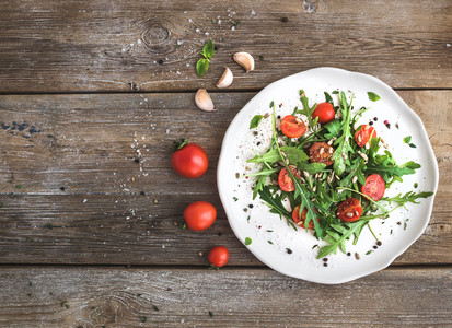 Salad with arugula  cherry tomatoes  sunflower seeds and herbs on white ceramic plate over rustic wood background  top view