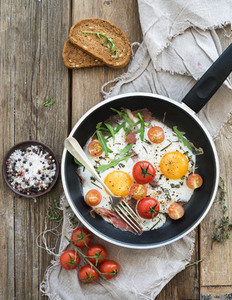 Pan of fried eggs  bacon and cherry tomatoes with bread on rustic wood table surface