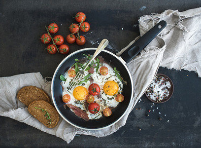 Pan of fried eggs bacon and cherry tomatoes with bread on dark table surface
