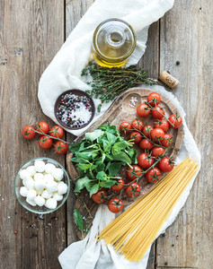 Ingredients for cooking pasta Spaghetti basil cherry tomatoes mozarella olive oil thyme salt spices on rustic chopping board over old wood background
