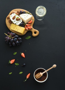 Cheese appetizer selection or whine snack set Variety of cheese grapes pecan nuts strawberry and honey on round wooden board over black backdrop top view