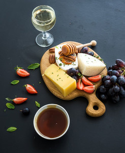 Cheese appetizer selection or whine snack set  Variety of cheese  grapes  pecan nuts  strawberry and honey on round wooden board over black backdrop