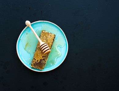 Honeycomb with honey dipper on blue ceramic plate over black background  top view