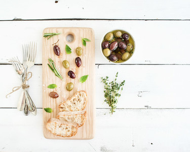 Mediterranean olives with herbs and ciabatta slices on rustic wooden board  over white background  top view