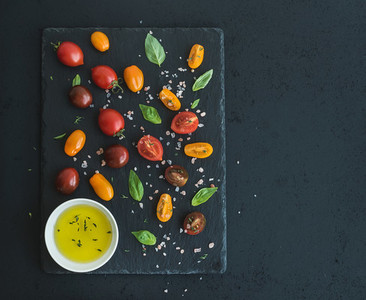 Cherry tomatoes of various color basil leaves spices and olive oil on black slate tray over dark grunge background Top view