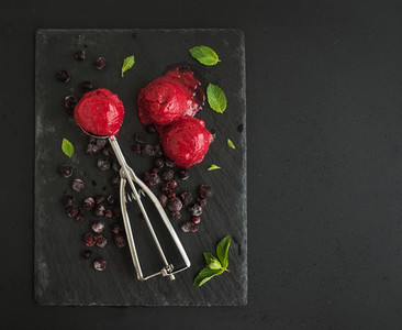Scoops of berry sorbet or ice cream with frozen black currant mint and metal scooper on black slate tray over dark grunge backdrop top view