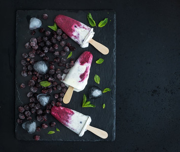 Black currant milk ice creams or popsicles with frozen black currant  ice cubes and mint on black slate tray over dark grunge backdrop  top view