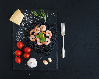 Black pasta spaghetti with shrimps  basil  pesto sauce  garlic  parmesan cheese and cherry tomatoes on a slate tray over black grunge backdrop  top view