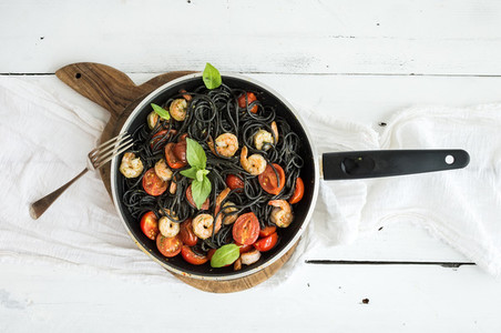 Black pasta spaghetti with shrimps  basil  pesto sauce and slow roasted cherry tomatoes in cooking pan on rustic chopping board over white wooden table