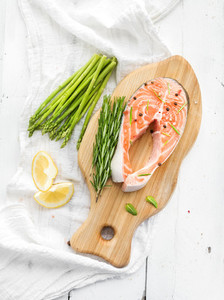 Raw salmon steak with asparagus  lemon  spices and rosemary on rustic wooden chopping board over white backdrop  Top view