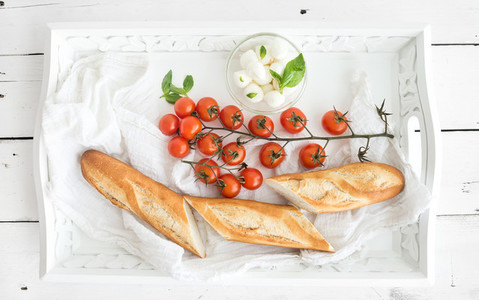 Baguette with branch of cherry tomatoes basil and mozzarella cheese on rustic white wooden tray top view