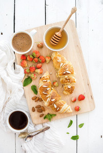 Freshly baked almond croissants with garden strawberries  mint  honey and coffee on serving board over white rustic wood backdrop