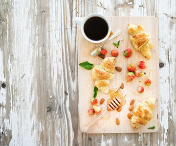 Freshly baked almond croissants with garden strawberries and honey on serving board over white rustic wood backdrop top view