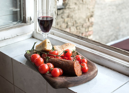 Home wine appetizer set Glass of red wine Hungarian spicy sausage farmers cheese and cherry tomatoes in a ructic wooden dish placed near window