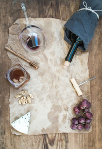 Wine and appetizer set with copy space in center  Glass of red wine  bottle  corkscrewer  blue cheese  grapes  honey  walnuts on oily craft paper over rustic wooden table  top view