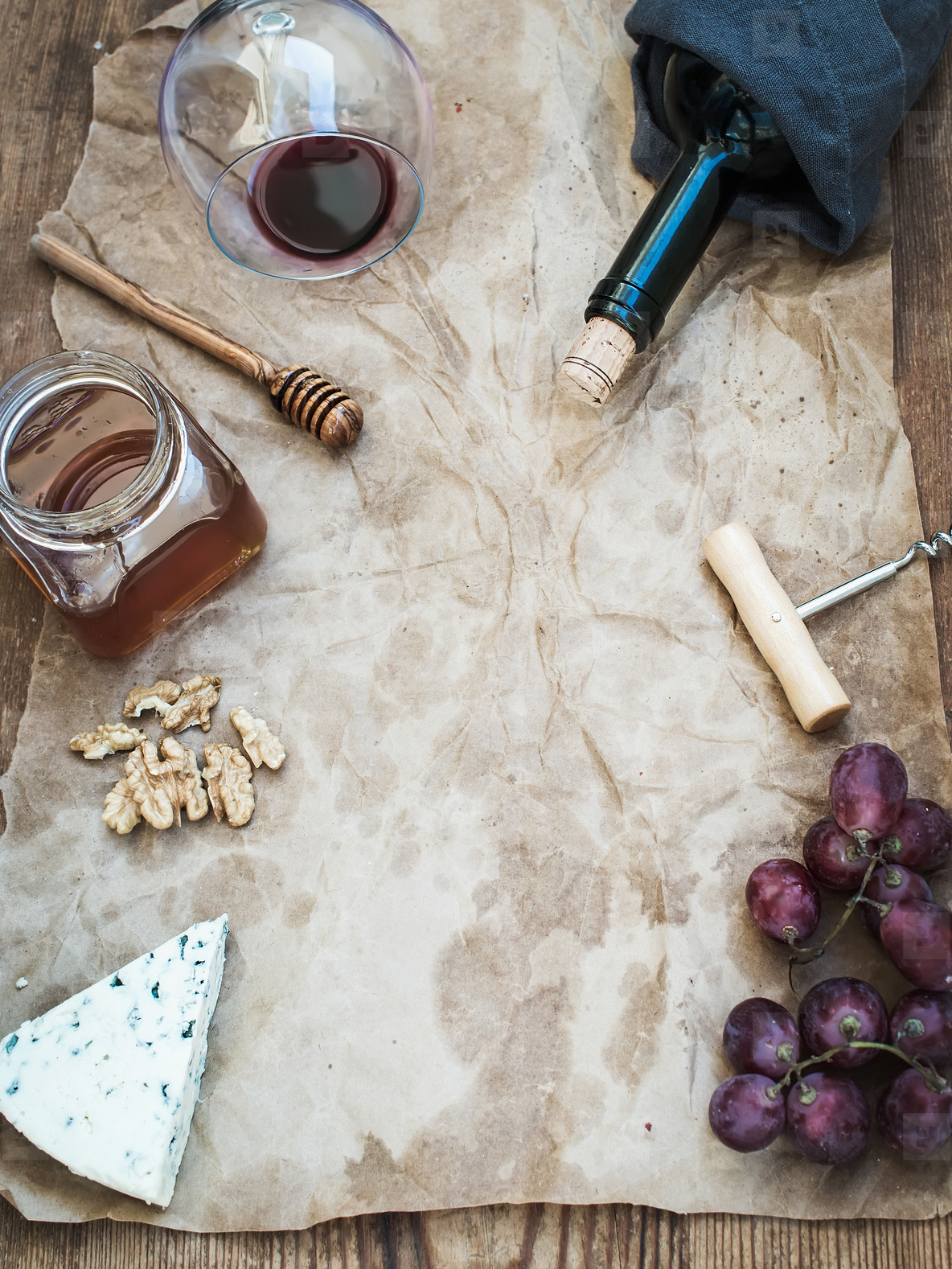 Wine and appetizer set with copy space in center. Glass of red wine, bottle, corkscrewer, blue cheese, grapes, honey, walnuts on oily craft paper over rustic wooden table, top view.