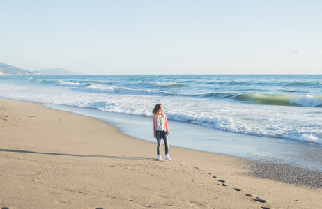 Young woman in red checkered shirt  jeans  white sneakers walking along beach and the stormy ocean on sunny winter day