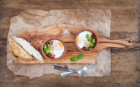 Country style breakfast set  Eggs baked in separate clay cups with tomatoes  peppers  fresh basil  bread slices on rustic board over oily craft paper and wooden background  top view