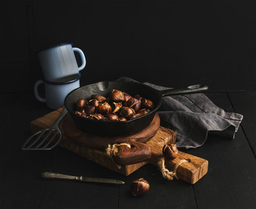 Roasted chestnuts in skillet cooking pan over rusti wooden boards  blue enamel mugs  towel on dark background