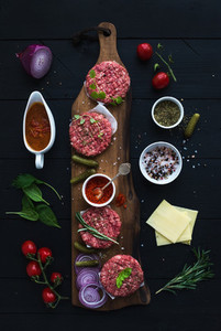 Ingredients for cooking burgers Raw ground beef meat cutlets on wooden chopping board red onion cherry tomatoes greens pickles tomato sauce cheese herbs and spices over black background top v
