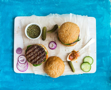 Fresh beef burger with cheese  vegetables  spicy tomato sauce on paper and white serving board  blue wooden background
