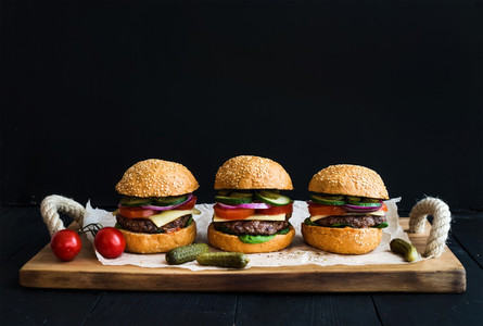 Fresh beef burgers with cheese  vegetables  pickles and spicy tomato sauce on paper over rustic wooden tray  black background