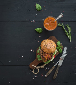Fresh homemade burger on dark serving board with spicy tomato sauce sea salt and herbs over black wooden background Top view