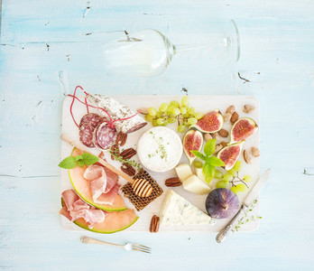 Wine and snack set  Figs  grapes  nuts  cheese variety  meat appetizers  herbs  glass on light blue background  top view