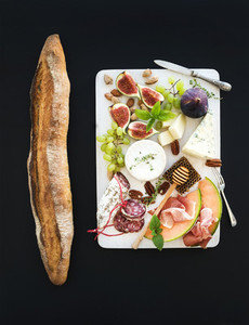Wine and snack set Baguette figs grapes nuts cheese variety meat appetizers herbs on white wooden board over black grunge background top view