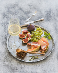 Wine appetizer set  Glass of white wine  honeycomb with drizzlier  figs  grapes  melon and prosciutto on silver tray over rustic grunge surface