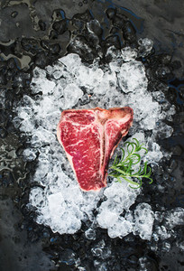 Raw fresh meat t bone steak on chipped ice with rosemary over dark slate stone backdrop