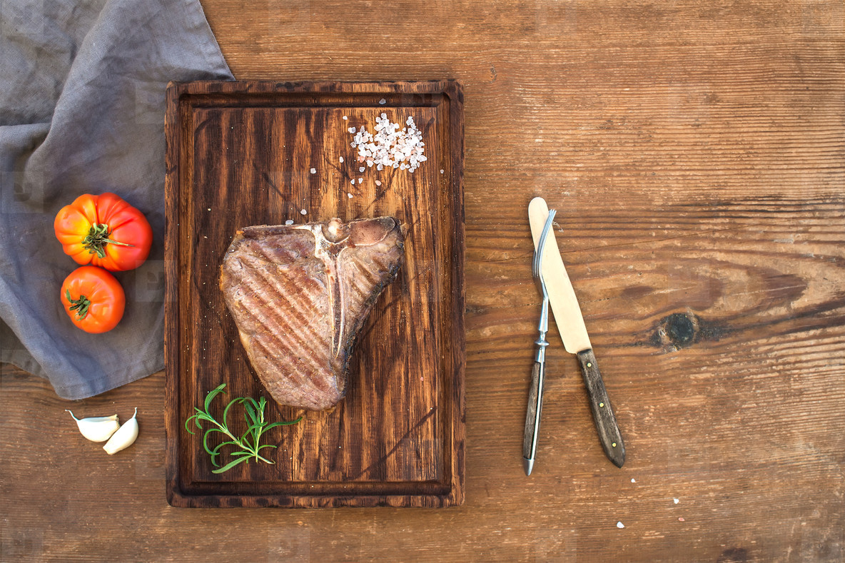 Cooked meat t bone steak on serving board with garlic cloves  tomatoes  rosemary and spices over rustic wooden background