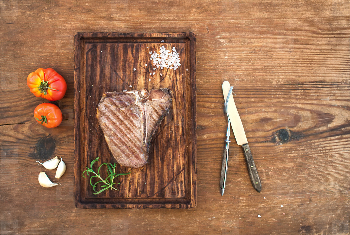 Cooked meat t-bone steak on serving board with garlic cloves, tomatoes, rosemary and spices over rustic wooden background.