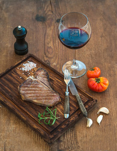 Cooked meat t bone steak on serving board with garlic cloves  tomatoes  rosemary  spices and glass of red wine over rustic wooden background