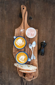 Homemade pumpkin cream soup in enamel mugs with herbs and fresh bread slices on olive serving board over rustic wooden background
