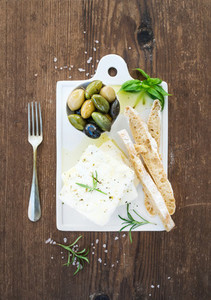 Fresh feta cheese with olives  basil  rosemary and bread slices on white ceramic serving board over rustic wooden background