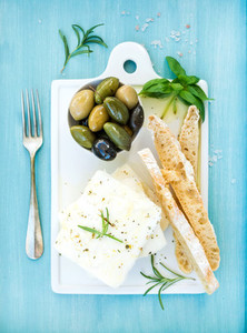 Fresh feta cheese with olives basil rosemary and bread slices on white ceramic serving board over bright turquoise blue painted wooden background