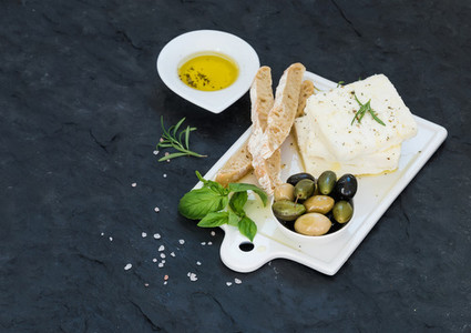 Fresh feta cheese with olives  basil  rosemary and bread slices on white ceramic serving board over black slate stone background