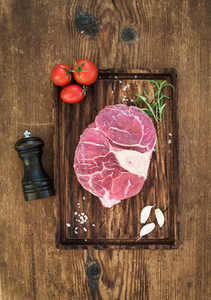 Raw fresh beef meat cross cut for ossobuco with garlic cloves  cherry tomatoes  rosemary  pepper and salt on serving board over rustic wooden background