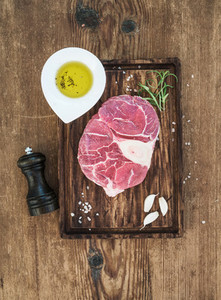 Raw fresh beef meat cross cut for ossobuco with garlic cloves  rosemary  pepper  oil and salt on serving board over rustic wooden background