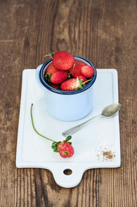 Fresh ripe red strawberries in blue enamel mug on white ceramic board over rustic wooden background