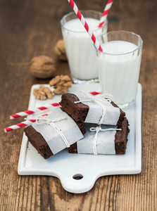 Chocolate brownie slices wrapped in paper and tired with rope  glasses of milk  stripe straws  walnuts on white ceramic board over rustic wooden background