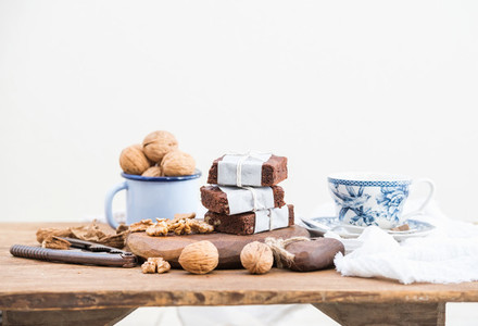 Chocolate brownie slices wrapped in paper and tired with rope  porcelain tea cup  enamel mug of walnuts on rustic wooden table  white background
