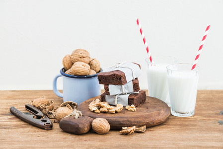 Chocolate brownie slices wrapped in paper and tired with rope milk glasses stripe straws enamel mug of walnuts on rustic wooden table white background
