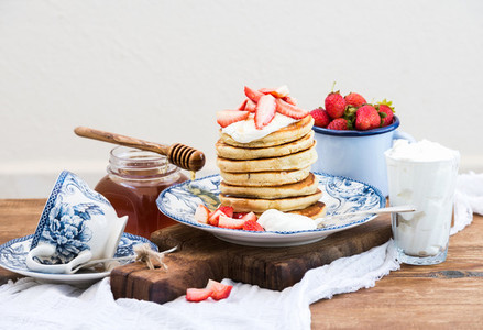 Breakfast set  Pancakes with fresh strawberries  sour cream and honey on a porcelain plate over rustic wooden table  Blue enamel cup full of berries behind  white background