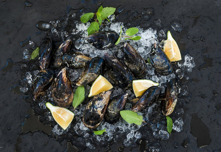 Fresh uncooked mussels with lemon  herbs and spices on chipped ice over dark slate stone backdrop