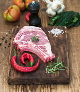 Raw fresh meat ribeye steak with salt chili pepper rosemary tomatoes garlic and spinach on rustic cutting board over wooden background
